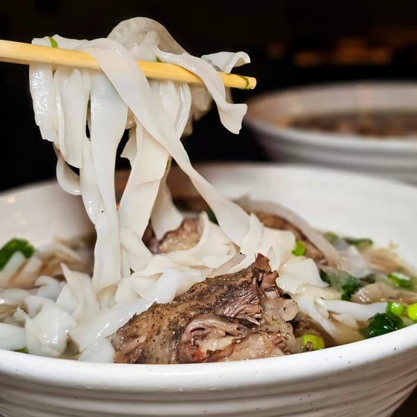 Chopsticks lifting wide rice noodles from a bowl of pho with meat and green onions.