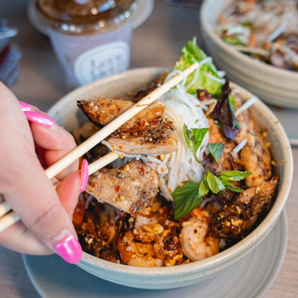 A hand with pink nails uses chopsticks to pick up grilled chicken from a Vietnamese noodle bowl.