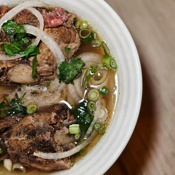 Close-up of a bowl of pho with beef, noodles, onions, and green herbs in broth.