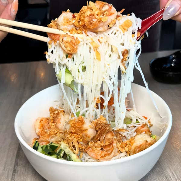 Chopsticks lifting a large portion of vermicelli noodles with shrimp and fried garlic from a white bowl.