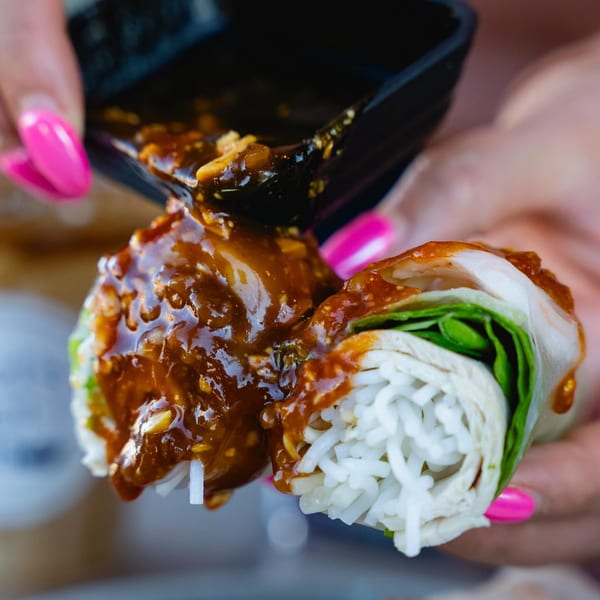 Hands with pink nails pour peanut sauce over a fresh Vietnamese spring roll.