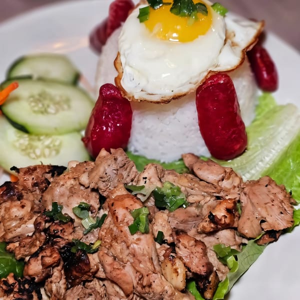 Vietnamese grilled chicken with rice, a fried egg, red sausages, and sliced cucumbers.